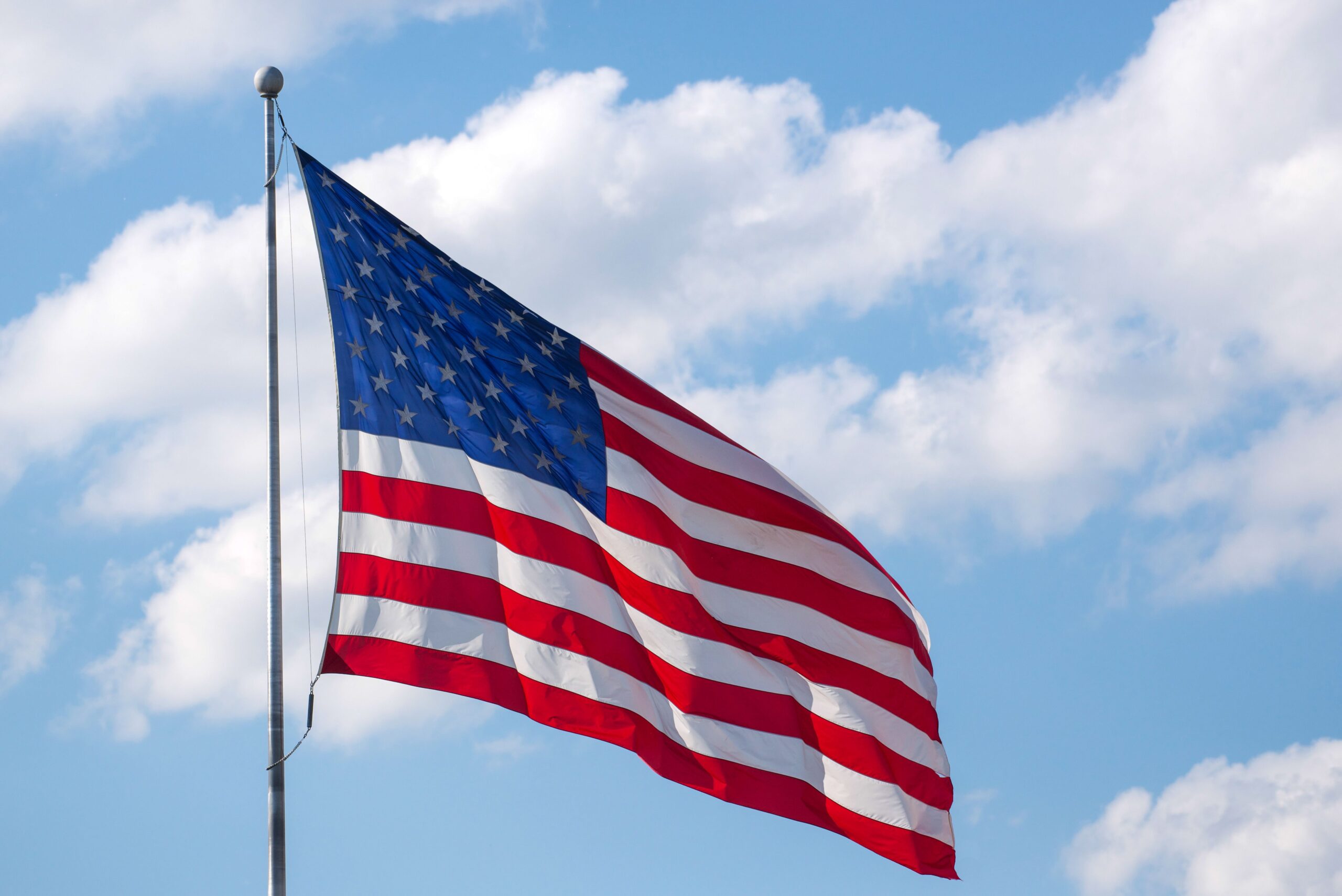 American flag waving against a clear blue sky with fluffy white clouds, symbolizing patriotism.